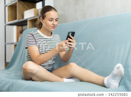 Teenage girl sits or lies on a sofa and scrolls through her smartphone. She chatting online Teenage girl sits or lies on a sofa and scrolls through her smartphone. She chatting online 142255081