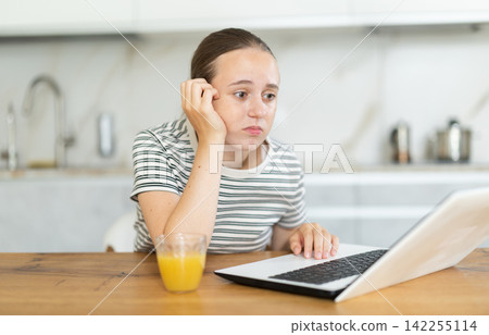 Teenage girl having problems with laptop while sitting at table in kitchen Teenage girl having problems with laptop while sitting at table in kitchen 142255114