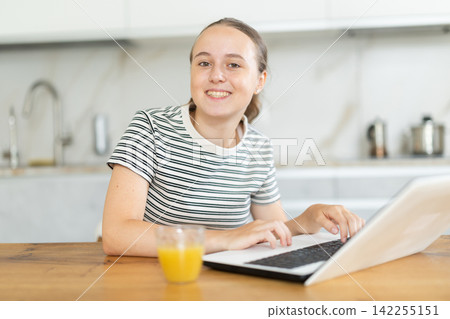 Teenage girl chatting on internet using laptop in domestic kitchen Teenage girl chatting on internet using laptop in domestic kitchen 142255151