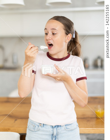Teenage girl eating fresh yogurt with spoon in modern kitchen Teenage girl eating fresh yogurt with spoon in modern kitchen 142255165
