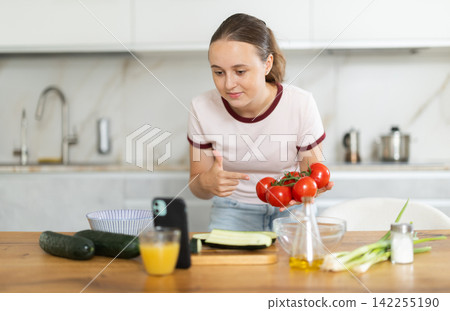 Teenage girl prepares salad in kitchen and looks at smartphone screen Teenage girl prepares salad in kitchen and looks at smartphone screen 142255190