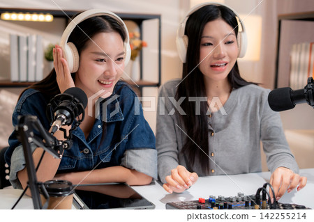 Two young asian woman with headphones speaking on microphone in home studio. Two young asian woman with headphones speaking on microphone in home studio. 142255214