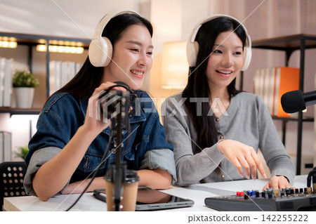 Two young asian woman with headphones speaking on microphone in home studio. Two young asian woman with headphones speaking on microphone in home studio. 142255222