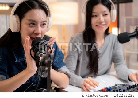 Two young asian woman with headphones speaking on microphone in home studio. Two young asian woman with headphones speaking on microphone in home studio. 142255223