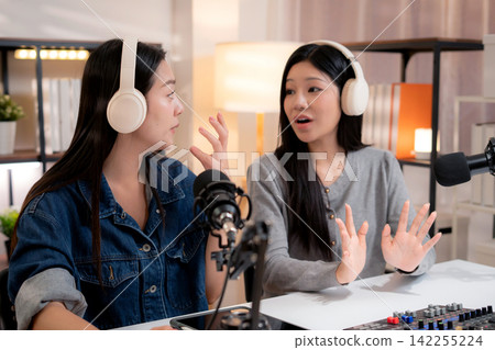 Two young asian woman with headphones speaking on microphone in home studio. Two young asian woman with headphones speaking on microphone in home studio. 142255224