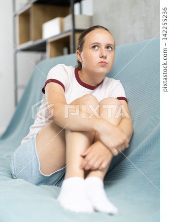 Uupset girl is sitting on couch in her room. She thinking and worrying about something Uupset girl is sitting on couch in her room. She thinking and worrying about something 142255236