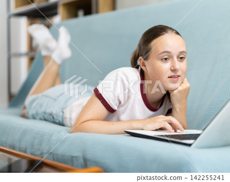 Teenage girl working on laptop while lying on cozy sofa Teenage girl working on laptop while lying on cozy sofa 142255241