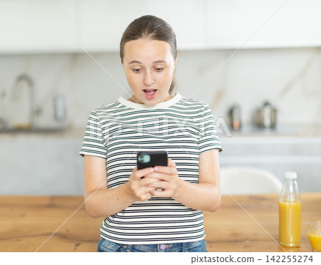 Girl scrolling through social media feed on mobile in kitchen Girl scrolling through social media feed on mobile in kitchen 142255274