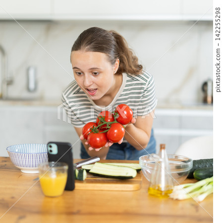 Positive teenage housewife using her smartphone during cooking at kitchen Positive teenage housewife using her smartphone during cooking at kitchen 142255298