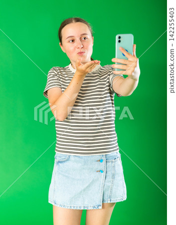 Happy teenage girl in denim skirt takes selfie on a smartphone on green background. Chroma key compositing Happy teenage girl in denim skirt takes selfie on a smartphone on green background. Chroma key compositing 142255403