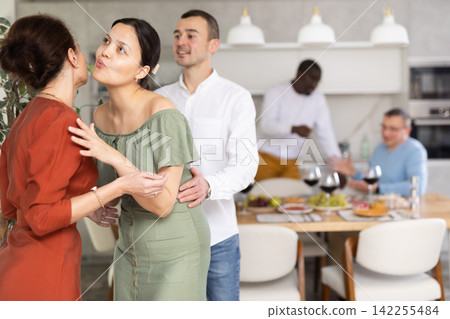 Women greet each other with kiss on cheek before festive dinner. Men shaking hands in background 142255484