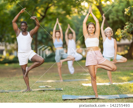 Multinational yoga enthusiasts with female coach perform vrikshasana in park Multinational yoga enthusiasts with female coach perform vrikshasana in park 142255493
