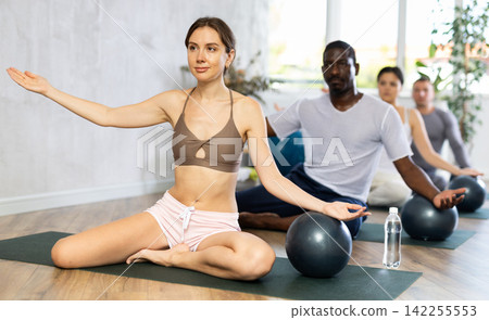 Woman in activewear sitting on yoga mat during group workout with fitness ball at gym. Healthy lifestyle concept Woman in activewear sitting on yoga mat during group workout with fitness ball at gym. Healthy lifestyle concept 142255553