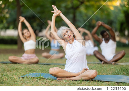Senior woman stretching in lotus during group yoga in park Senior woman stretching in lotus during group yoga in park 142255600