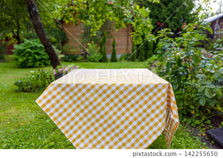 Empty Table with White and Yellow Checkered Cotton Tablecloth in Summer Garden 142255650