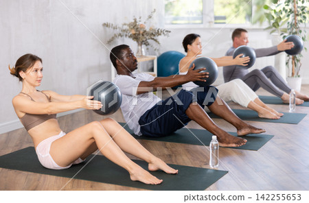 Woman in activewear sitting on yoga mat during group workout with fitness ball at gym. Healthy lifestyle concept Woman in activewear sitting on yoga mat during group workout with fitness ball at gym. Healthy lifestyle concept 142255653