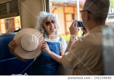 Asian senior elderly man taking a photo of his wife with camera inside a vintage train carriage. Asian senior elderly man taking a photo of his wife with camera inside a vintage train carriage. 142256680
