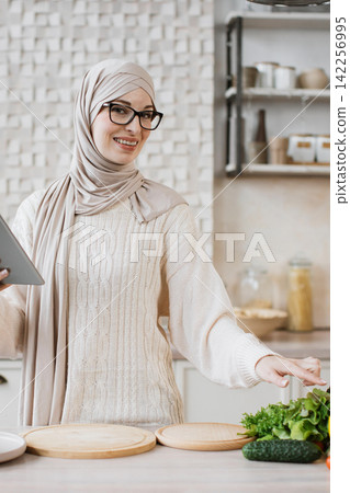 A smiling woman wearing a hijab and glasses is in a kitchen, preparing a healthy meal with fresh vegetables and a tablet A smiling woman wearing a hijab and glasses is in a kitchen, preparing a healthy meal with fresh vegetables and a tablet 142256995