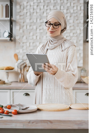 A smiling woman wearing glasses and a hijab is holding a tablet in a modern kitchen, looking at the screen while preparing food A smiling woman wearing glasses and a hijab is holding a tablet in a modern kitchen, looking at the screen while preparing food 142256998