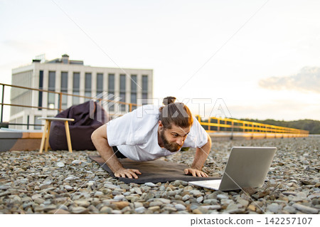 A man with a beard and bun hairstyle performs pushups on a yoga mat on a rooftop while watching a laptop A man with a beard and bun hairstyle performs pushups on a yoga mat on a rooftop while watching a laptop 142257107