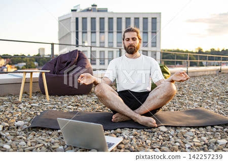 A man with a beard practices yoga and meditation on a rooftop terrace, using a laptop for guidance while sitting on a mat surrounded by pebbles A man with a beard practices yoga and meditation on a rooftop terrace, using a laptop for guidance while sitting on a mat surrounded by pebbles 142257239