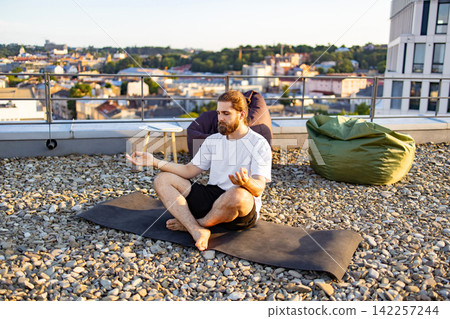 A man with a beard and tied-back hair sits in a lotus position on a yoga mat on a gravel rooftop, practicing meditation with a city skyline in the background A man with a beard and tied-back hair sits in a lotus position on a yoga mat on a gravel rooftop, practicing meditation with a city skyline in the background 142257244