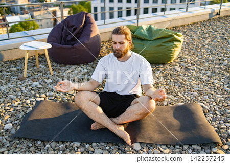 A man with a beard sits in a lotus position on a yoga mat on a gravel rooftop, practicing mindfulness with beanbag chairs and city buildings in the background A man with a beard sits in a lotus position on a yoga mat on a gravel rooftop, practicing mindfulness with beanbag chairs and city buildings in the background 142257245