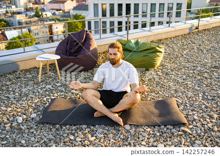 A man with a beard practices yoga and meditation on a rooftop terrace with beanbag chairs and a city view A man with a beard practices yoga and meditation on a rooftop terrace with beanbag chairs and a city view 142257246