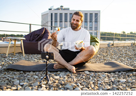 A bearded man sits cross-legged on a yoga mat on a rooftop, eating a salad and recording a video on his smartphone A bearded man sits cross-legged on a yoga mat on a rooftop, eating a salad and recording a video on his smartphone 142257252