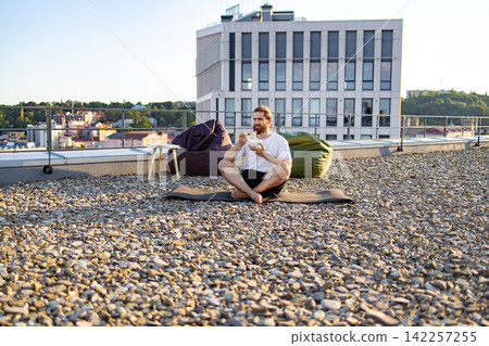 A man with a beard sits cross-legged on a yoga mat on a gravel rooftop terrace, eating from a bowl while beanbag chairs and a modern building are visible in the background A man with a beard sits cross-legged on a yoga mat on a gravel rooftop terrace, eating from a bowl while beanbag chairs and a modern building are visible in the background 142257255