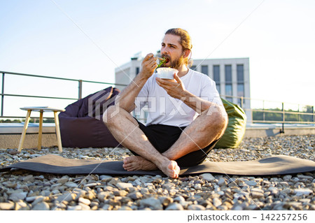 A man with a beard and tied-back hair sits cross-legged on a yoga mat on a gravel rooftop, eating a salad from a plastic container A man with a beard and tied-back hair sits cross-legged on a yoga mat on a gravel rooftop, eating a salad from a plastic container 142257256