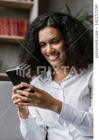 A young woman with curly hair smiles while looking at her smartphone, seated on a sofa with bookshelves in the background A young woman with curly hair smiles while looking at her smartphone, seated on a sofa with bookshelves in the background 142257290