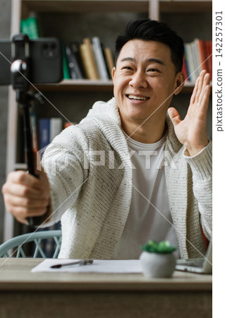 A smiling man is recording a video with his smartphone on a gimbal while sitting at a desk with a laptop 142257301