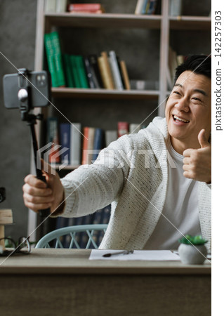 A smiling man with a thumbs-up gesture records a video on his smartphone using a selfie stick while sitting at a desk with a laptop A smiling man with a thumbs-up gesture records a video on his smartphone using a selfie stick while sitting at a desk with a laptop 142257303
