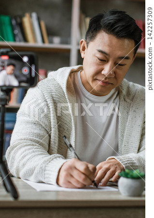 A man is intently studying and writing notes while a smartphone records him, suggesting online learning or content creation 142257309