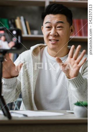A man is actively recording himself on a smartphone placed on a tripod, gesturing with his hands while sitting at a desk with a laptop A man is actively recording himself on a smartphone placed on a tripod, gesturing with his hands while sitting at a desk with a laptop 142257313