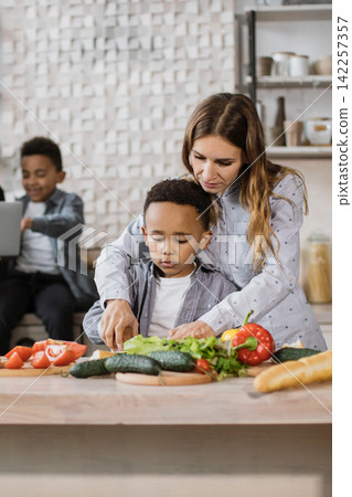 A mother and her two sons are preparing a healthy meal together in a bright, modern kitchen, chopping fresh vegetables for a salad A mother and her two sons are preparing a healthy meal together in a bright, modern kitchen, chopping fresh vegetables for a salad 142257357