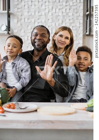 A diverse family with parents and two children smiling and waving hello from a kitchen setting A diverse family with parents and two children smiling and waving hello from a kitchen setting 142257395