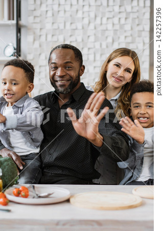 A diverse family with two children and parents smiling and waving at the camera in a bright, modern kitchen setting A diverse family with two children and parents smiling and waving at the camera in a bright, modern kitchen setting 142257396