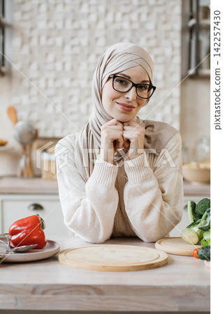 A smiling woman wearing a hijab and glasses is in a kitchen, ready to prepare a healthy meal with fresh vegetables A smiling woman wearing a hijab and glasses is in a kitchen, ready to prepare a healthy meal with fresh vegetables 142257430