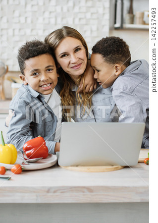 A mother and her two sons are gathered around a laptop in the kitchen, smiling and preparing food with fresh vegetables A mother and her two sons are gathered around a laptop in the kitchen, smiling and preparing food with fresh vegetables 142257493