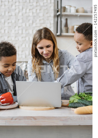 A mother and her two sons are gathered around a laptop in the kitchen, looking at recipes and preparing ingredients for a meal A mother and her two sons are gathered around a laptop in the kitchen, looking at recipes and preparing ingredients for a meal 142257495