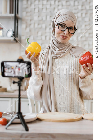A woman wearing a hijab and glasses records a cooking tutorial on her smartphone, holding colorful bell peppers A woman wearing a hijab and glasses records a cooking tutorial on her smartphone, holding colorful bell peppers 142257606