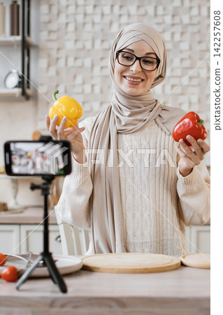 A smiling woman wearing a hijab and glasses holds a yellow and a red bell pepper while recording a video on her smartphone A smiling woman wearing a hijab and glasses holds a yellow and a red bell pepper while recording a video on her smartphone 142257608