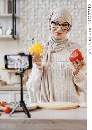A woman wearing a hijab and glasses records herself in a kitchen while holding bell peppers for a cooking demonstration A woman wearing a hijab and glasses records herself in a kitchen while holding bell peppers for a cooking demonstration 142257610