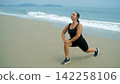 Young woman in black athletic wear performing a deep lunge stretch on a wide sandy beach with open ocean visible in background. Outdoor fitness concept 142258106