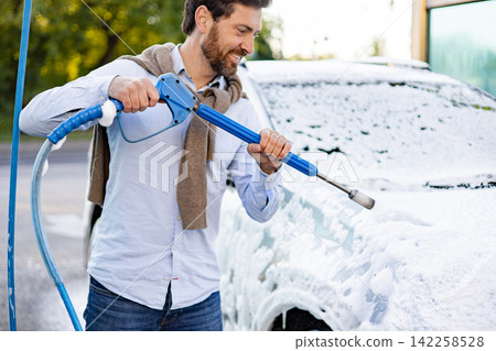 A smiling Caucasian man in jeans and a light blue shirt is using a high-pressure washer to apply foam to his car at a car wash A smiling Caucasian man in jeans and a light blue shirt is using a high-pressure washer to apply foam to his car at a car wash 142258528