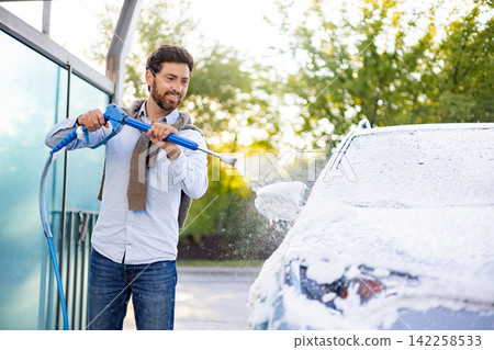 A smiling caucasian man is actively washing his car using a high-pressure water jet and foam at an outdoor car wash station A smiling caucasian man is actively washing his car using a high-pressure water jet and foam at an outdoor car wash station 142258533