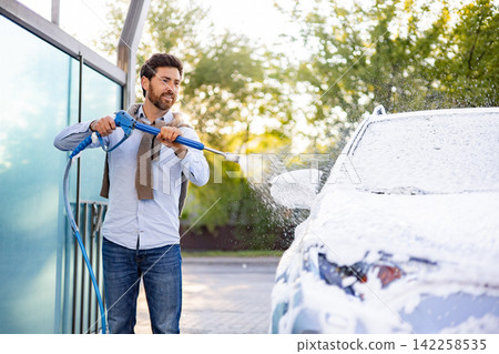 A smiling man with a beard is washing his car with a high-pressure washer, covering it in foam A smiling man with a beard is washing his car with a high-pressure washer, covering it in foam 142258535