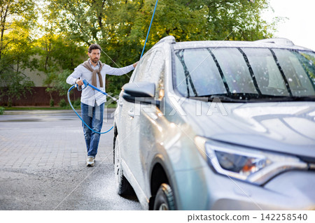 A caucasian man is actively washing his car using a high-pressure water hose outdoors on a sunny day A caucasian man is actively washing his car using a high-pressure water hose outdoors on a sunny day 142258540